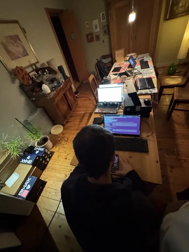 A developer seen from behind at a long table with several laptops, a receipt printer and boxed mini PCs, testing the full till setup